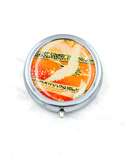 Round silver metal pill box featuring a white flying crane on an orange background with metallic gold details.