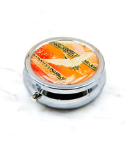 Round silver metal pill box featuring a white flying crane on an orange background with metallic gold details.