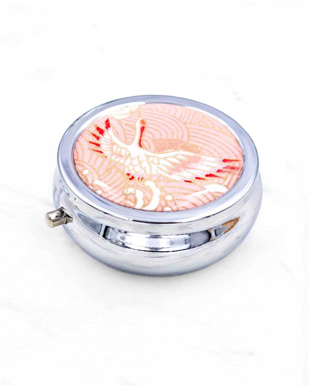 Silver metal pill box featuring a flying crane on a pink background with metallic gold accent.