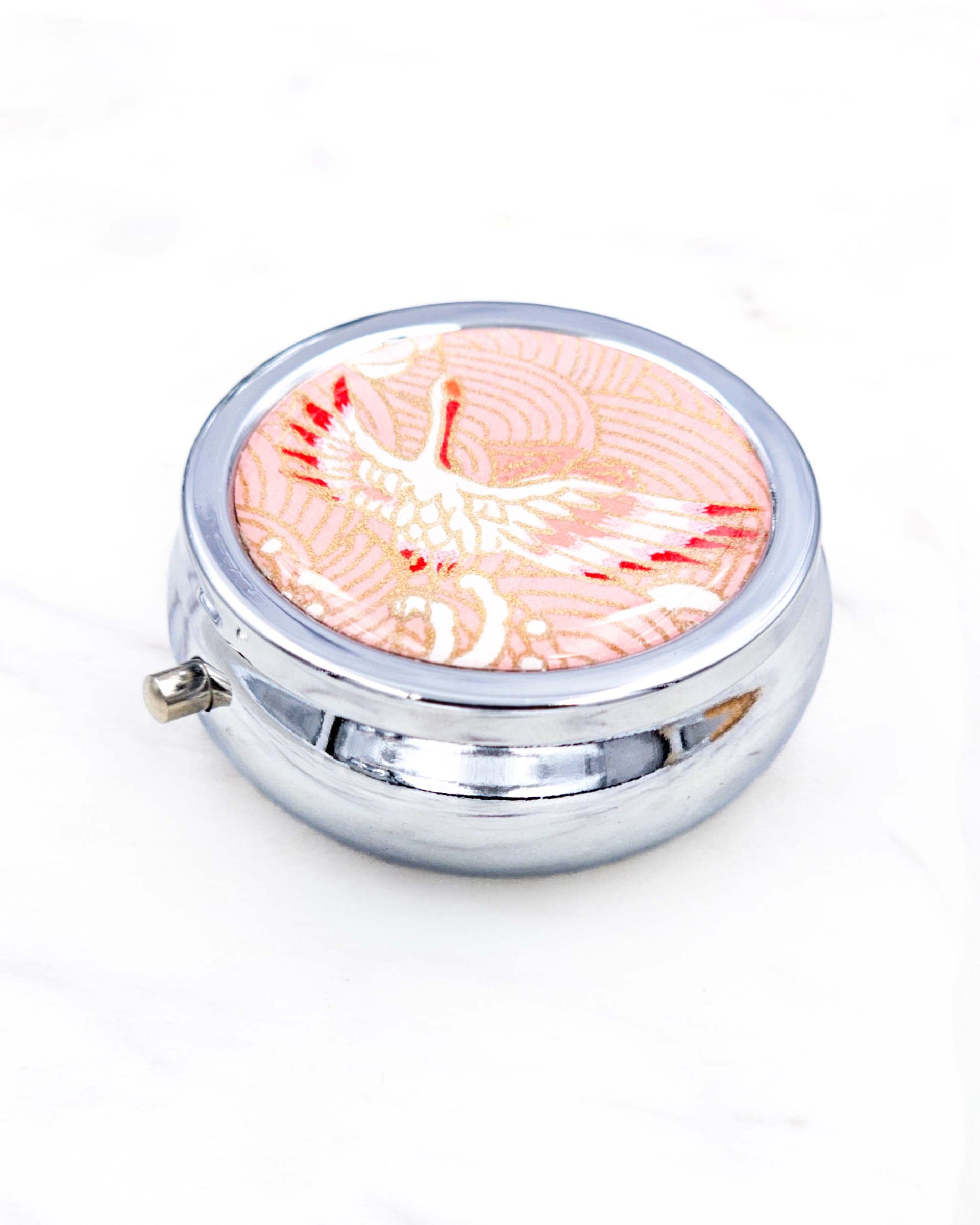Silver metal pill box featuring a flying crane on a pink background with metallic gold accent.