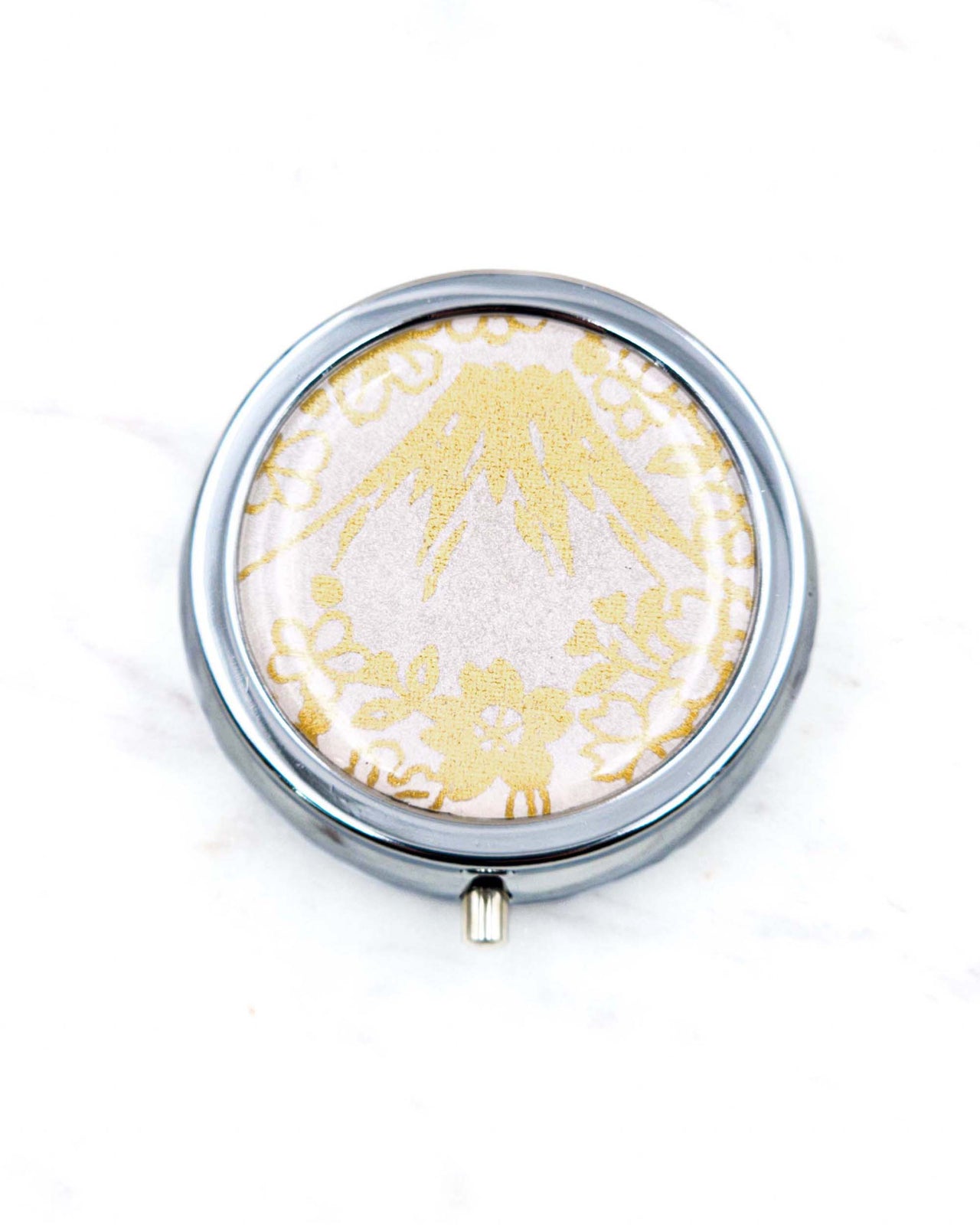 Round silver metal pill box featuring metallic gold Mt Fuji and cherry blossoms on a light pink background.