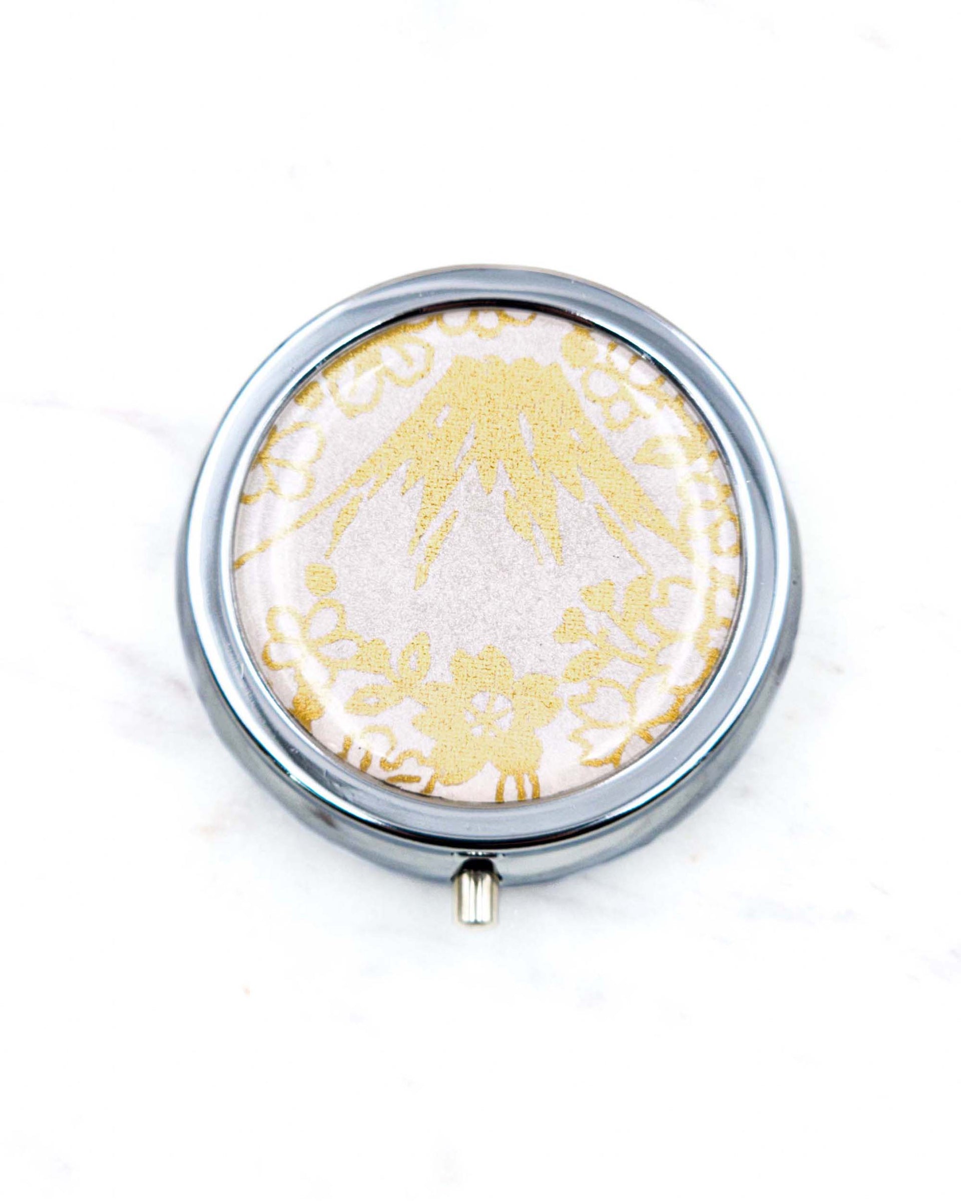 Round silver metal pill box featuring metallic gold Mt Fuji and cherry blossoms on a light pink background.