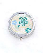Silver metal pill box featuring green, white, and blue cherry blossoms on green background with metallic gold accents.