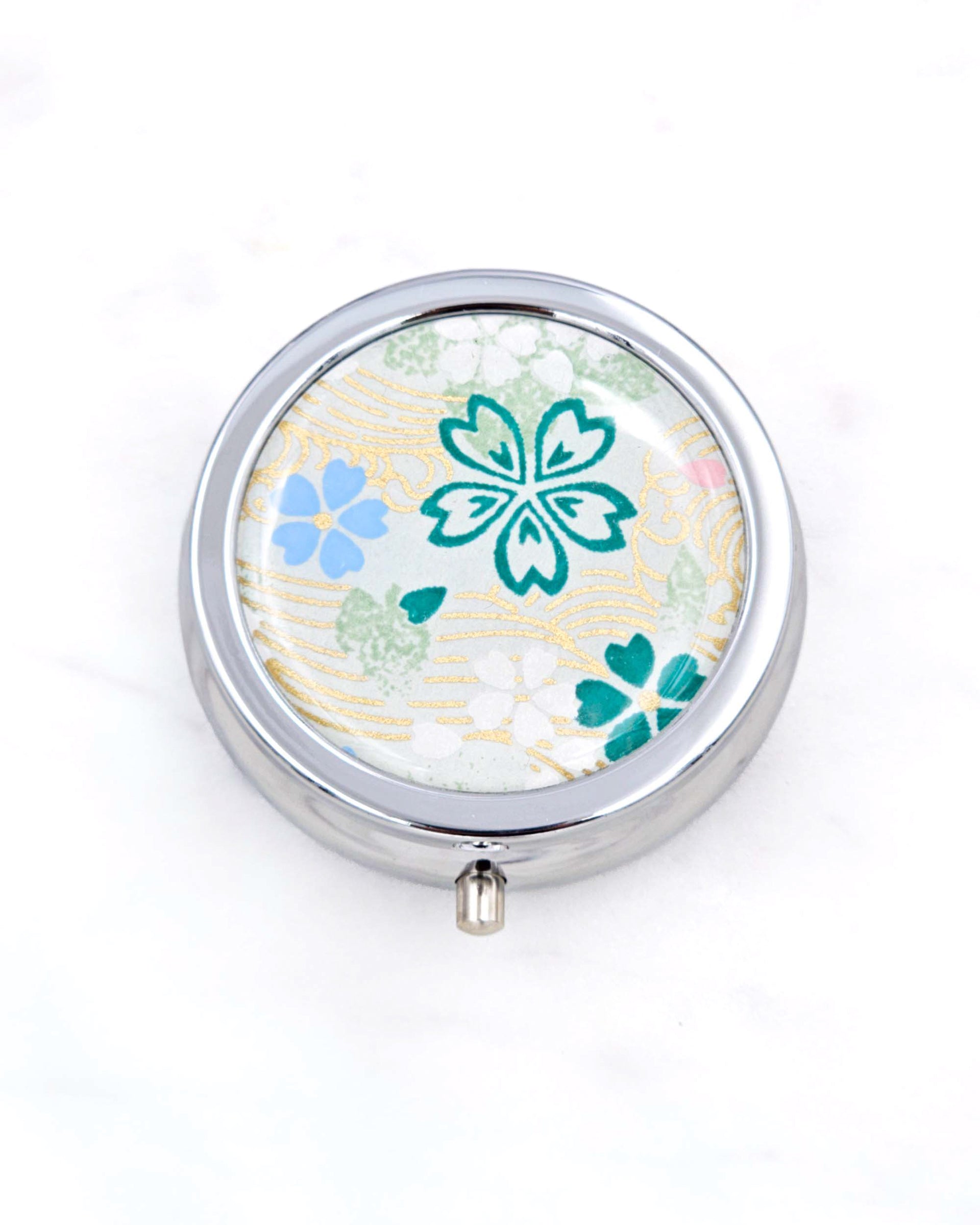 Silver metal pill box featuring green, white, and blue cherry blossoms on green background with metallic gold accents.