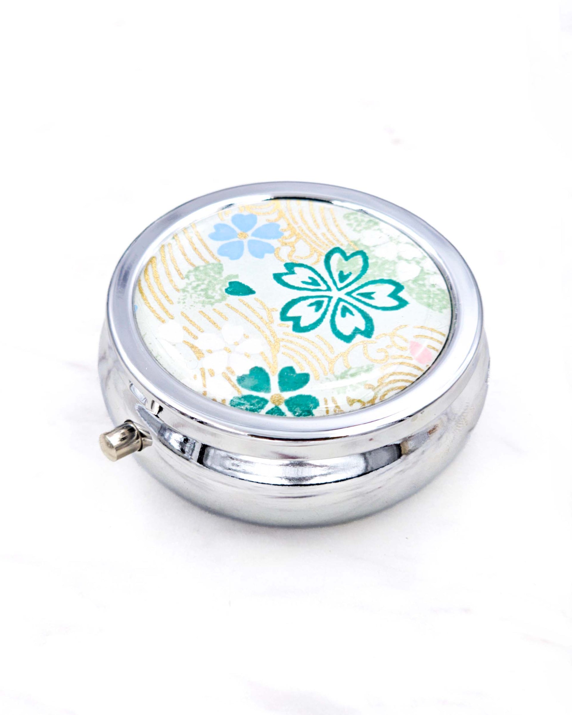 Silver metal pill box featuring green, white, and blue cherry blossoms on green background with metallic gold accents.