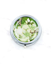 Round silver metal pill box featuring green and white cherry blossoms on a green background with metallic gold details.