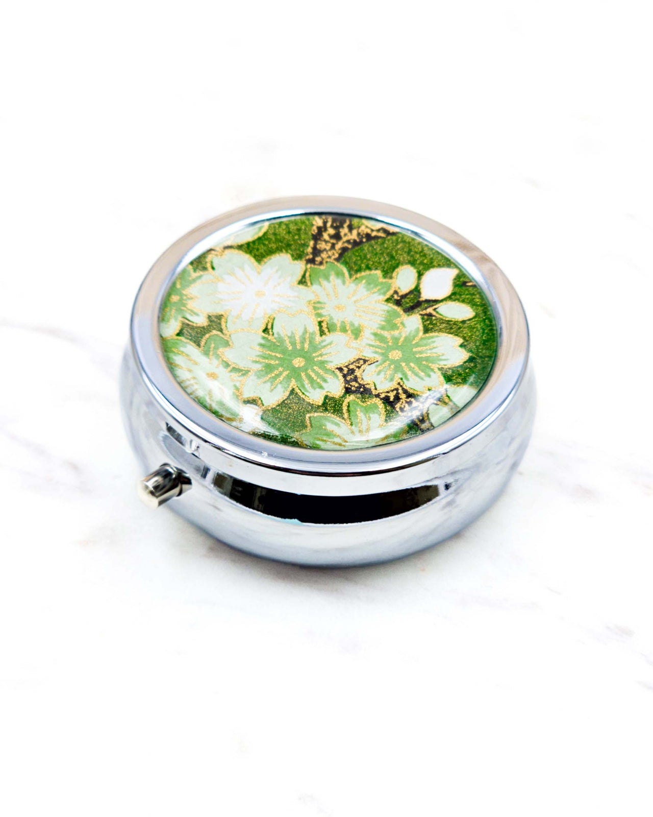 Round silver metal pill box featuring green and white cherry blossoms on a green background with metallic gold details.
