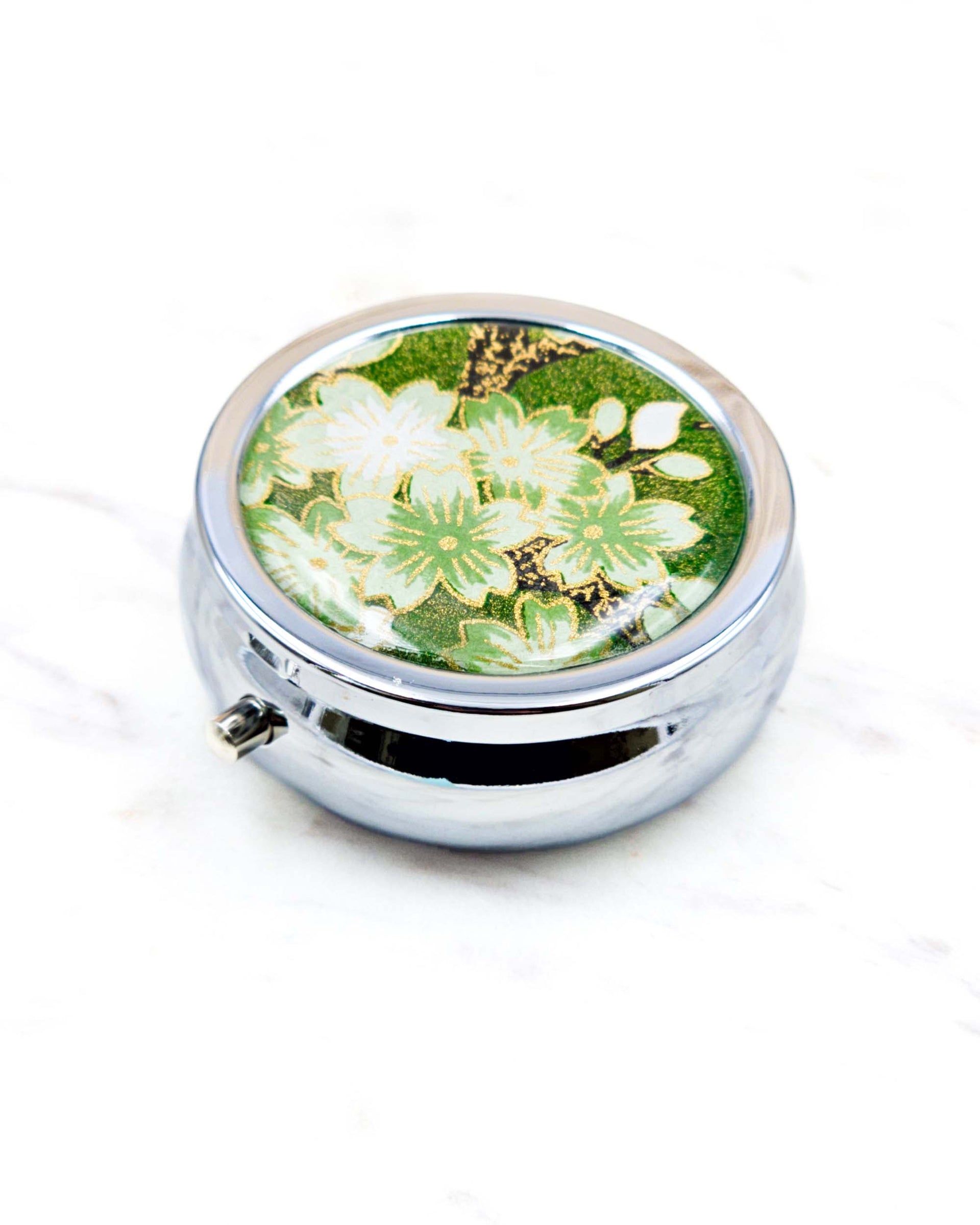 Round silver metal pill box featuring green and white cherry blossoms on a green background with metallic gold details.