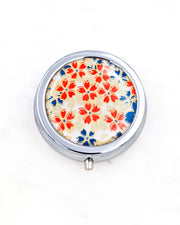 Silver metal pill box featuring red, white, and blue cherry blossoms on a white background with metallic gold accents.