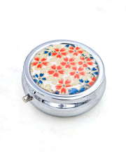 Silver metal pill box featuring red, white, and blue cherry blossoms on a white background with metallic gold accents.