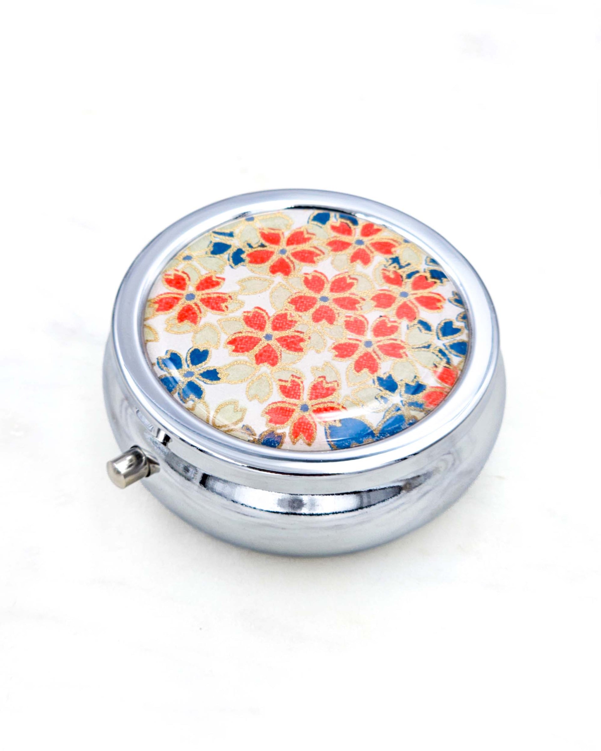 Silver metal pill box featuring red, white, and blue cherry blossoms on a white background with metallic gold accents.
