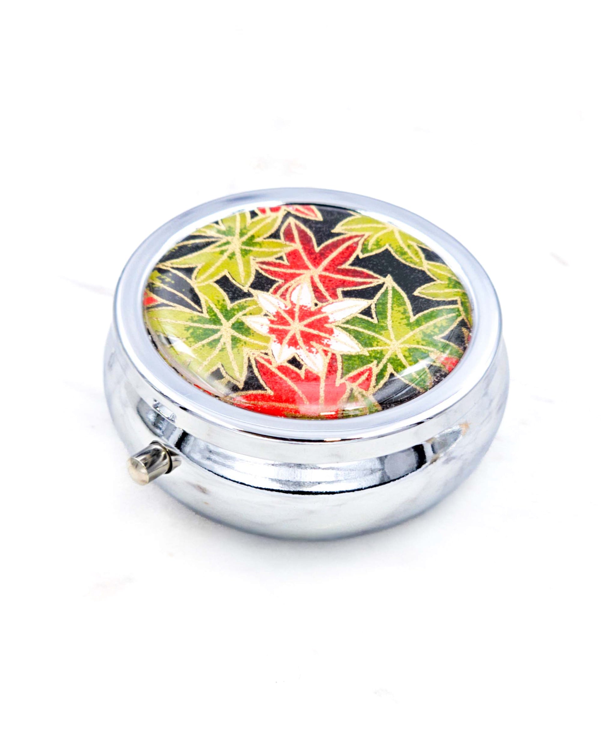 Silver metal pill box featuring green, red, and white maples leaves on a black background with metallic gold accents.