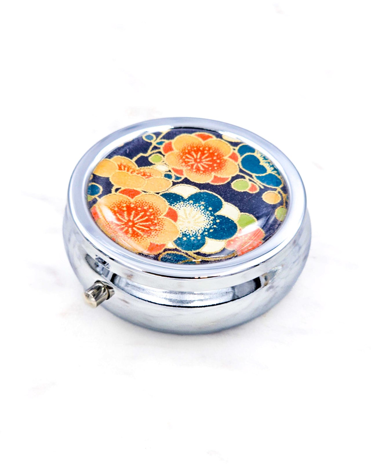 Silver metal pill box featuring orange and blue plum blossoms on a dark blue background with metallic gold accents.