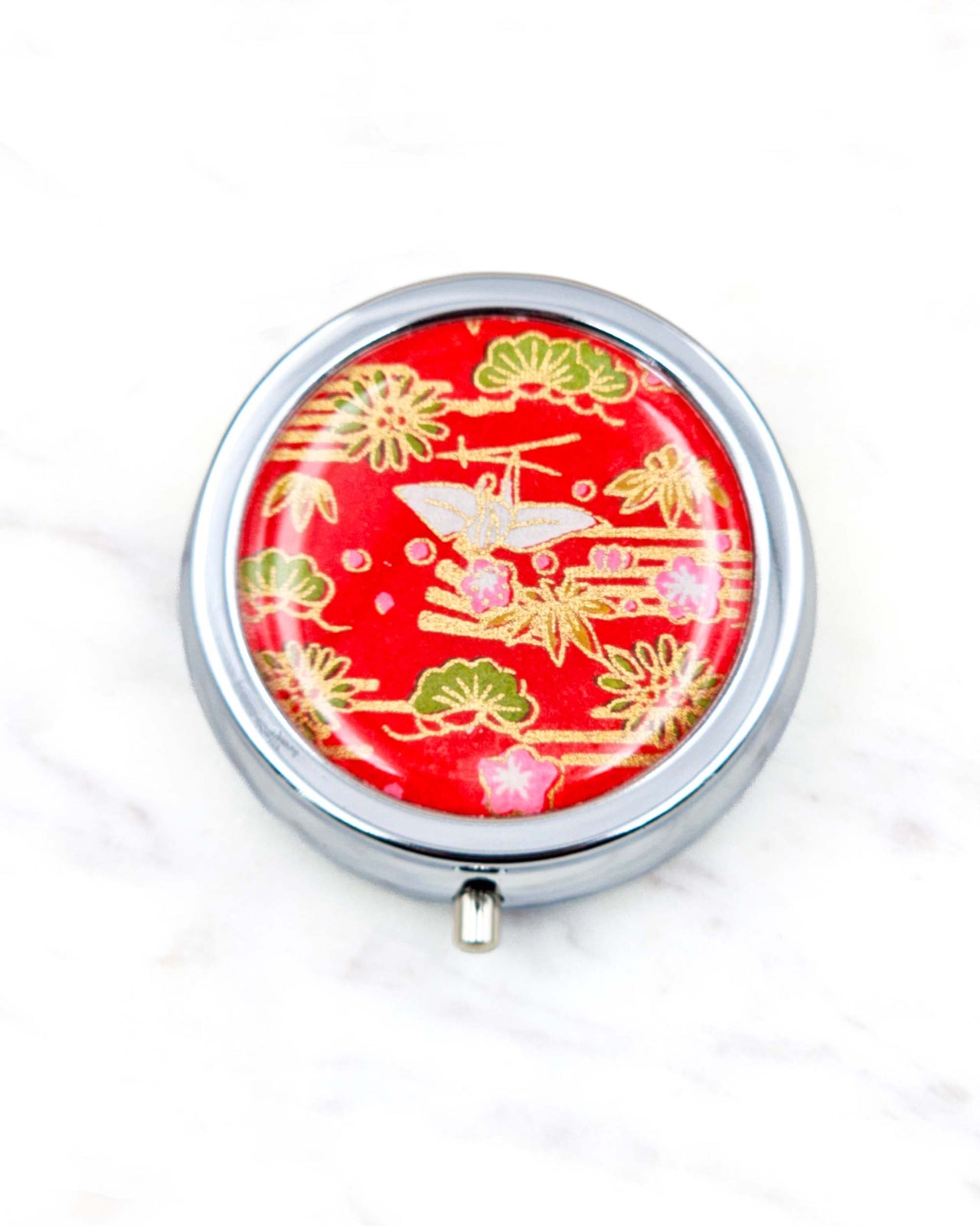 Round silver metal pill box featuring a white origami crane with green plants and pink plum blossoms on a red background with metallic gold details.