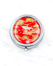 Round silver metal pill box featuring a white origami crane with green plants and pink plum blossoms on a red background with metallic gold details.