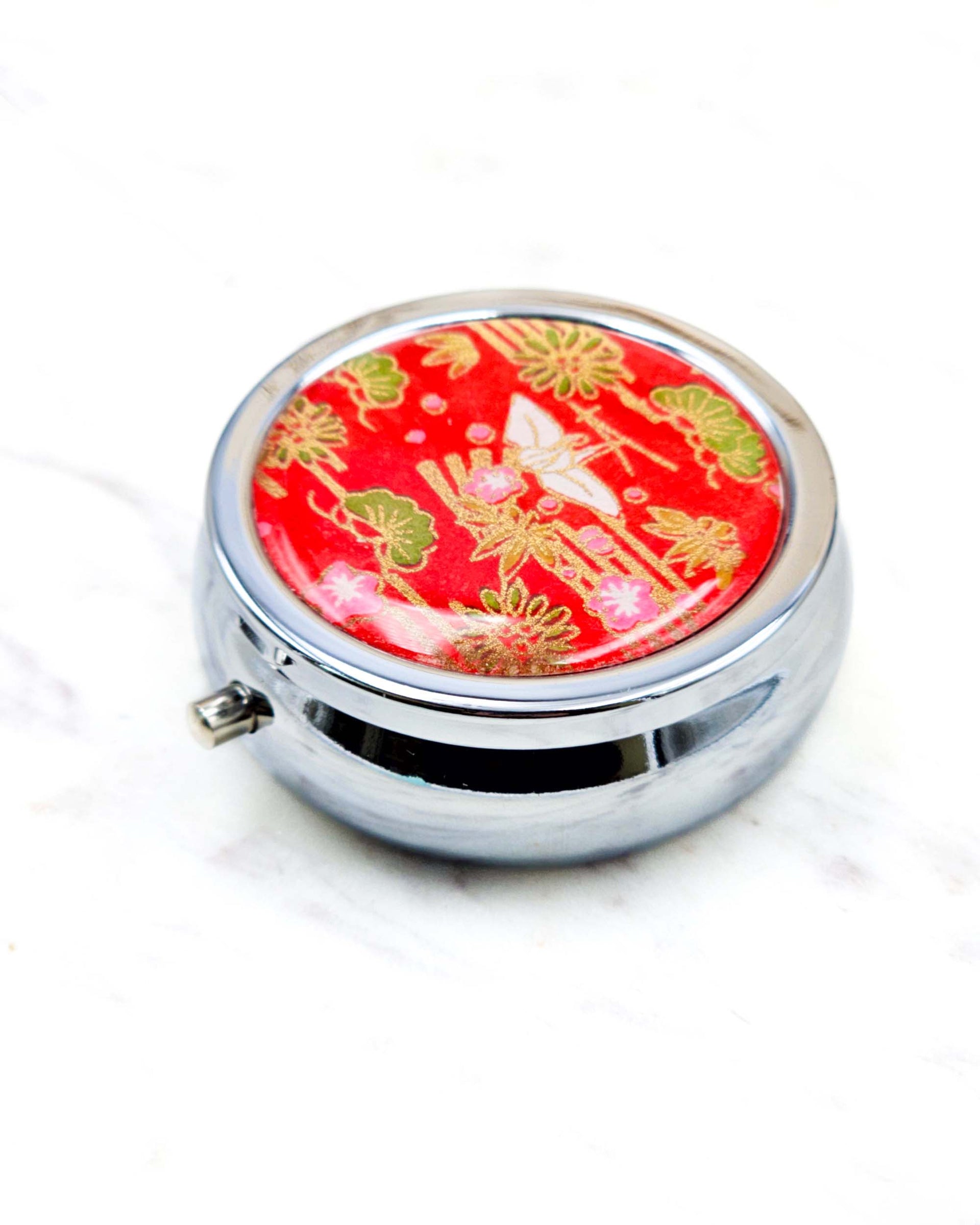 Round silver metal pill box featuring a white origami crane with green plants and pink plum blossoms on a red background with metallic gold details.