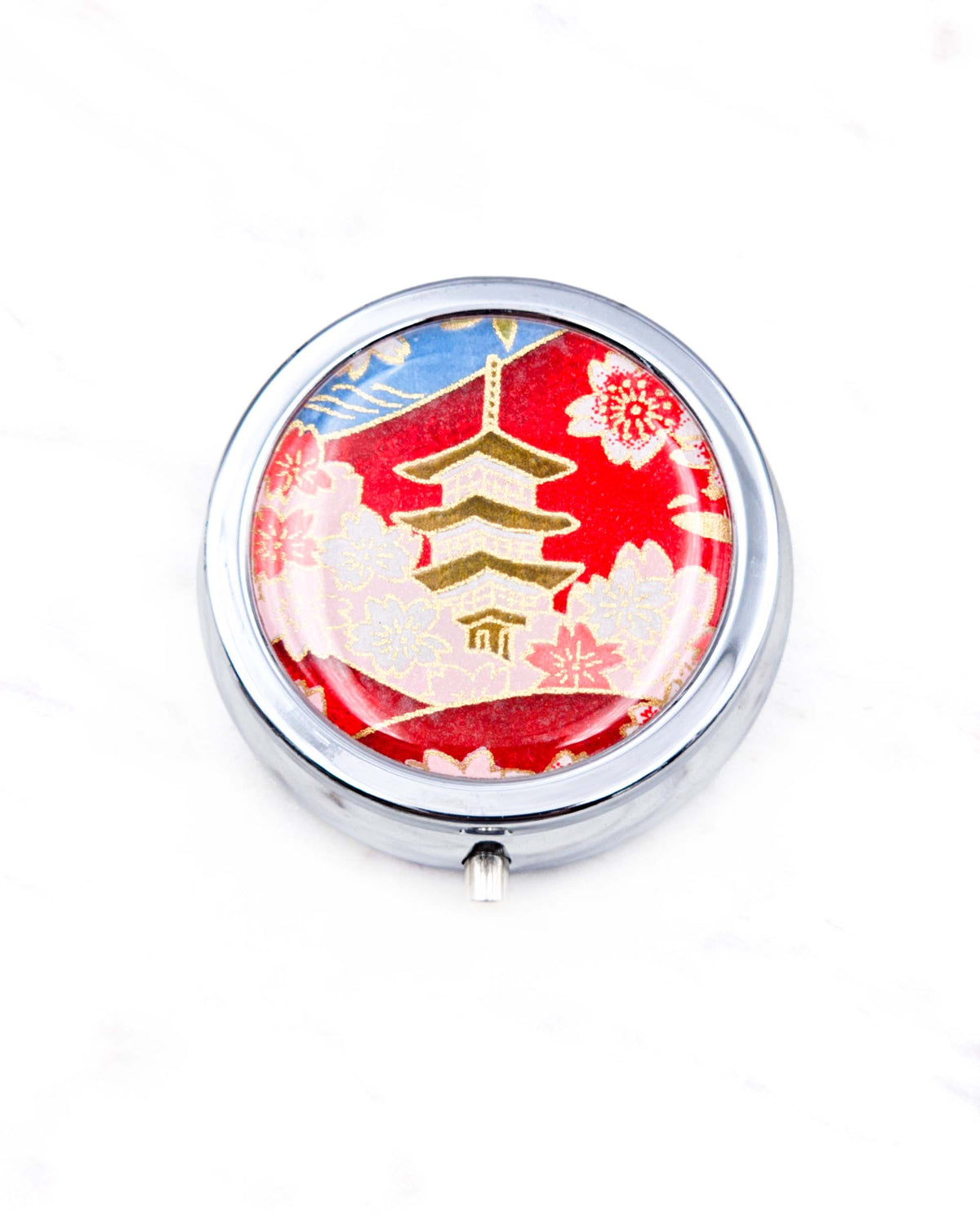Round silver metal pill box featuring a pagoda with pink, white, and red cherry blossoms on a red and blue background with metallic gold detail.