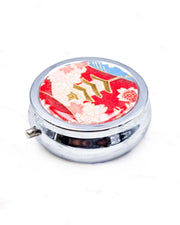 Round silver metal pill box featuring a pagoda with pink, white, and red cherry blossoms on a red and blue background with metallic gold detail.