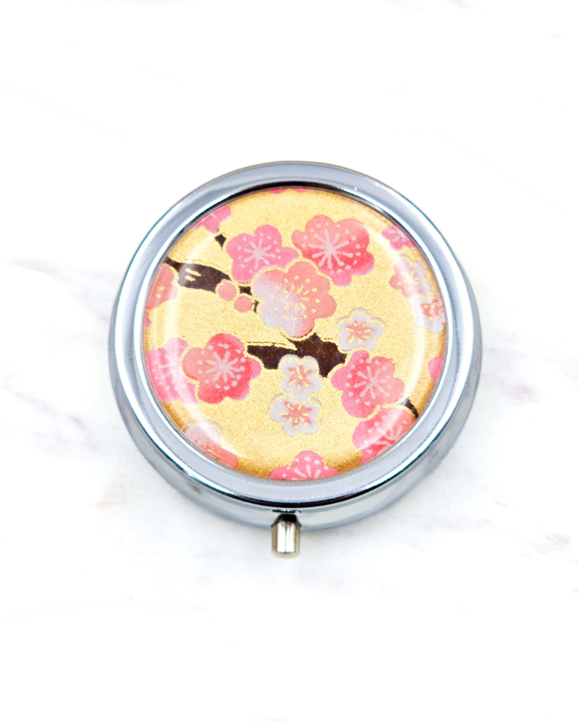 Round silver metal pill box featuring pink and white plum blossoms on a metallic gold background.