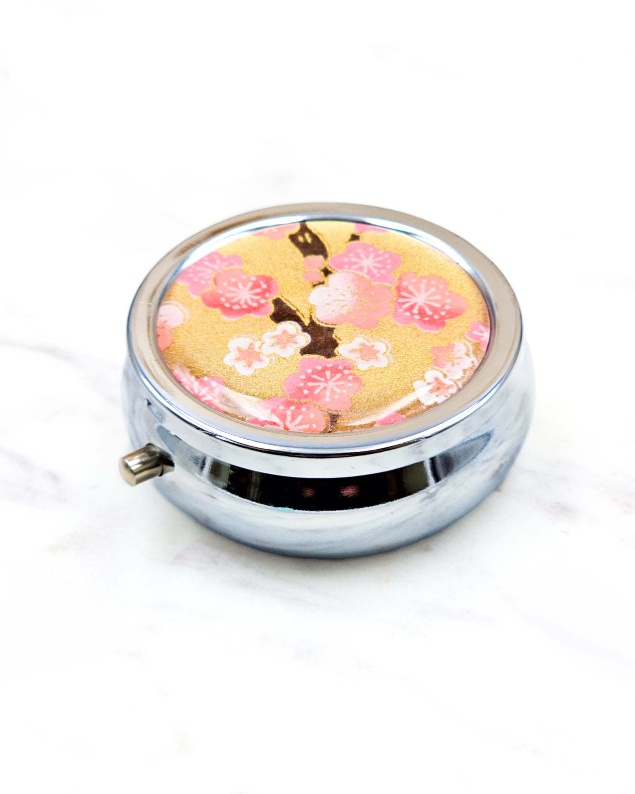 Round silver metal pill box featuring pink and white plum blossoms on a metallic gold background.