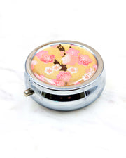 Round silver metal pill box featuring pink and white plum blossoms on a metallic gold background.