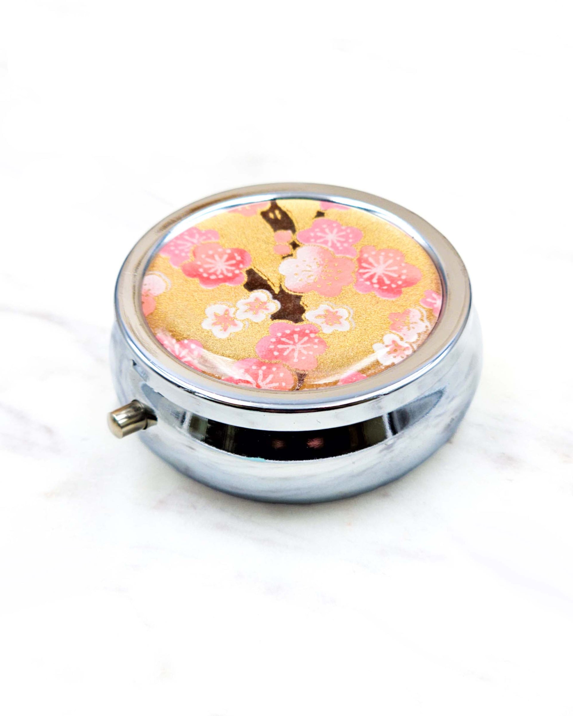 Round silver metal pill box featuring pink and white plum blossoms on a metallic gold background.