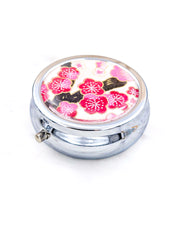 Silver metal pill box featuring a pink plum blossom tree on a white background with metallic gold accents.