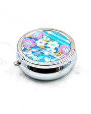 Round silver metal pill box featuring a white and pink cherry blossoms on a light blue background with metallic gold details.