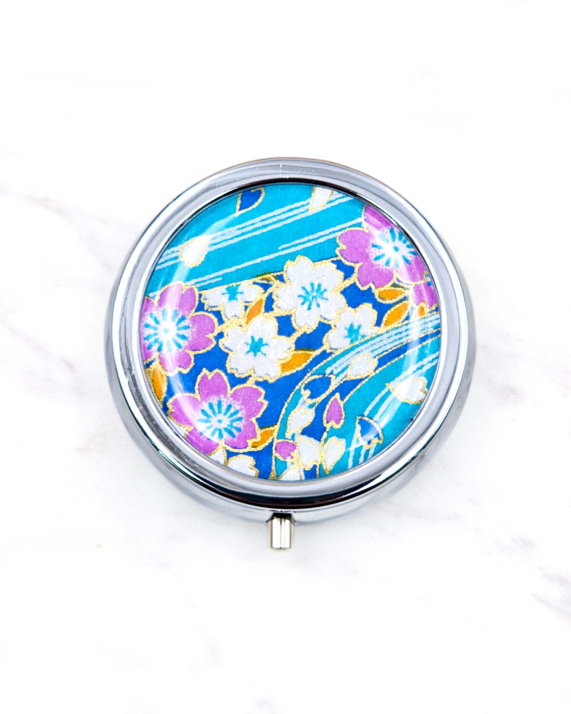 Round silver metal pill box featuring a white and pink cherry blossoms on a light blue background with metallic gold details.