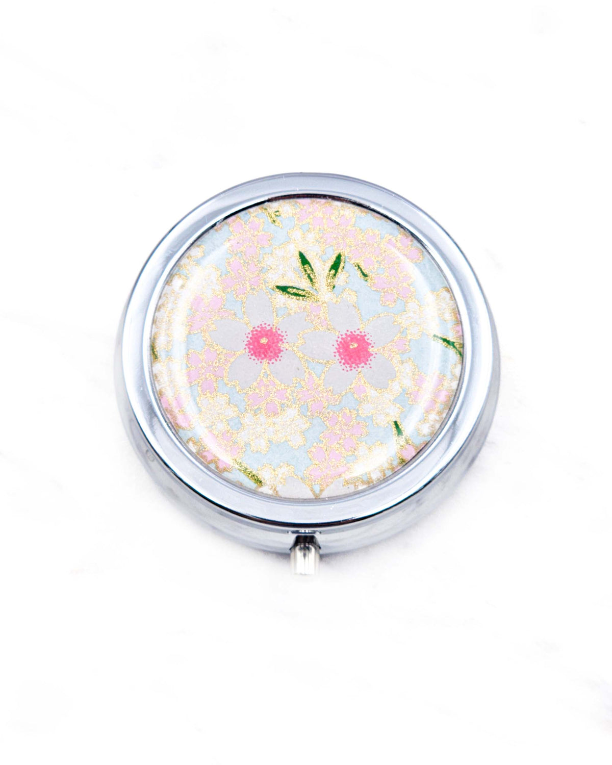 Round silver metal pill box featuring pink and white cherry blossoms on a light blue background with metallic gold details.