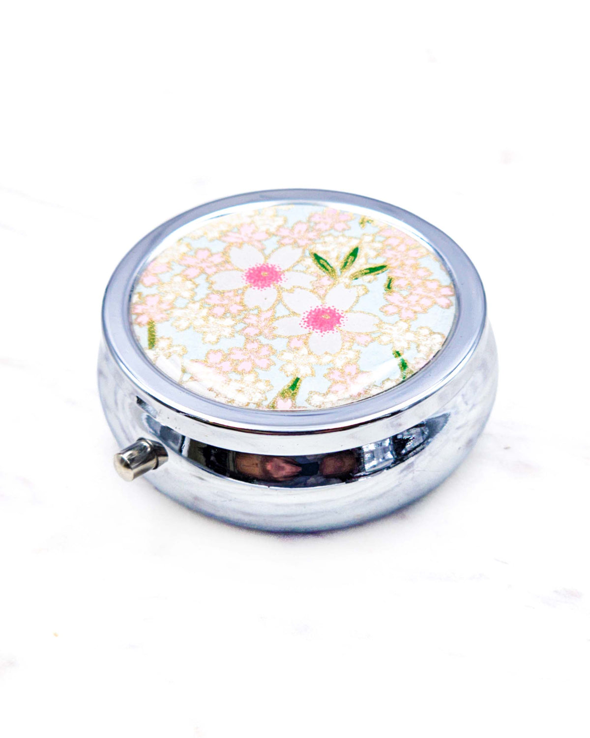 Round silver metal pill box featuring pink and white cherry blossoms on a light blue background with metallic gold details.