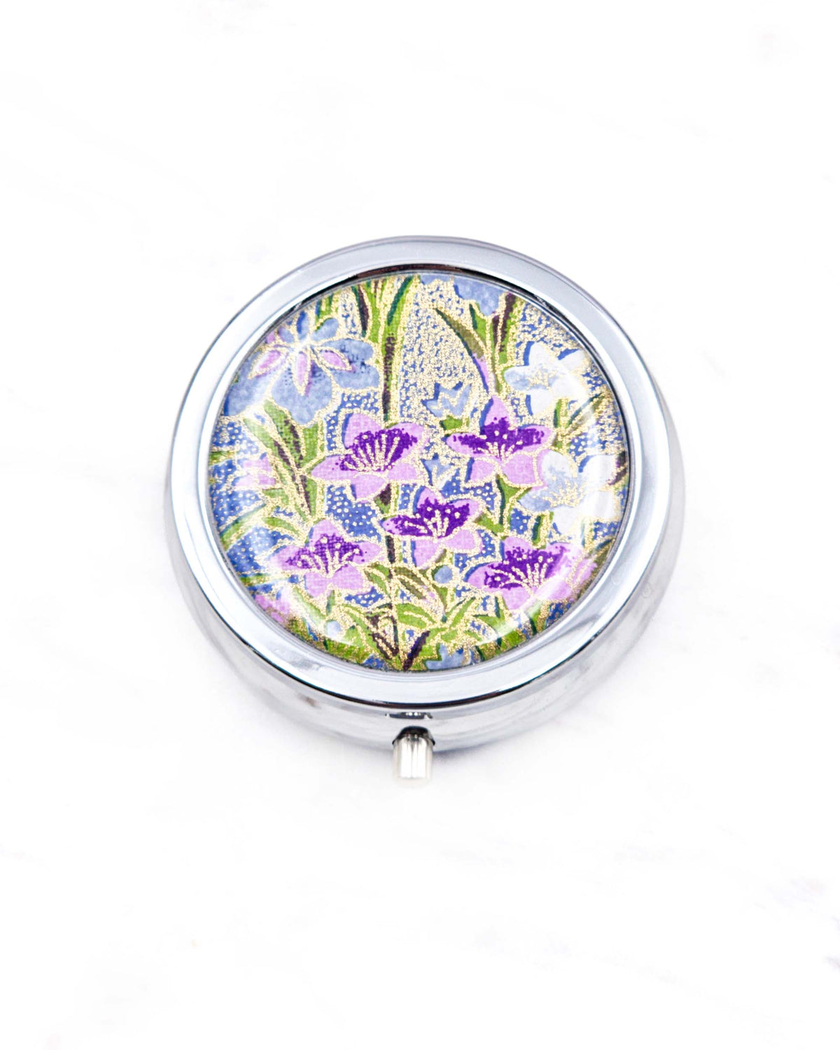 Round silver metal pill box featuring blue, purple, and white irises with green leaves and metallic gold details on a royal blue background.