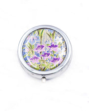 Round silver metal pill box featuring blue, purple, and white irises with green leaves and metallic gold details on a royal blue background.