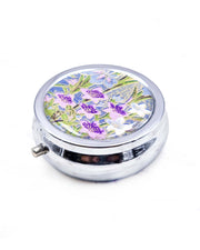 Round silver metal pill box featuring blue, purple, and white irises with green leaves and metallic gold details on a royal blue background.