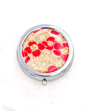 Silver metal pill box featuring red cherry blossoms on a brown background with metallic gold accents.