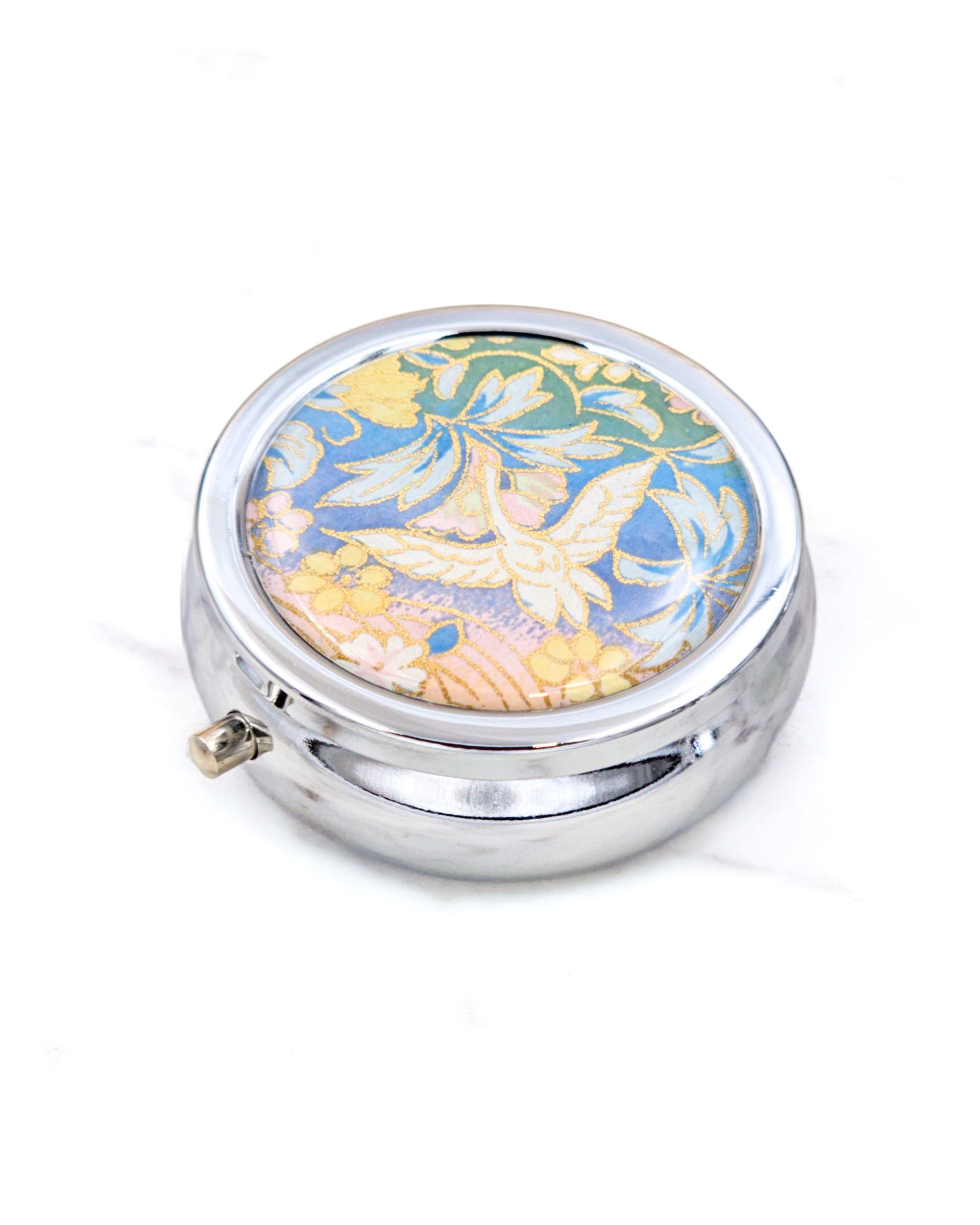 Silver metal pill box featuring a white flying crane on a green, blue , and pink pastel background with metallic gold accents.