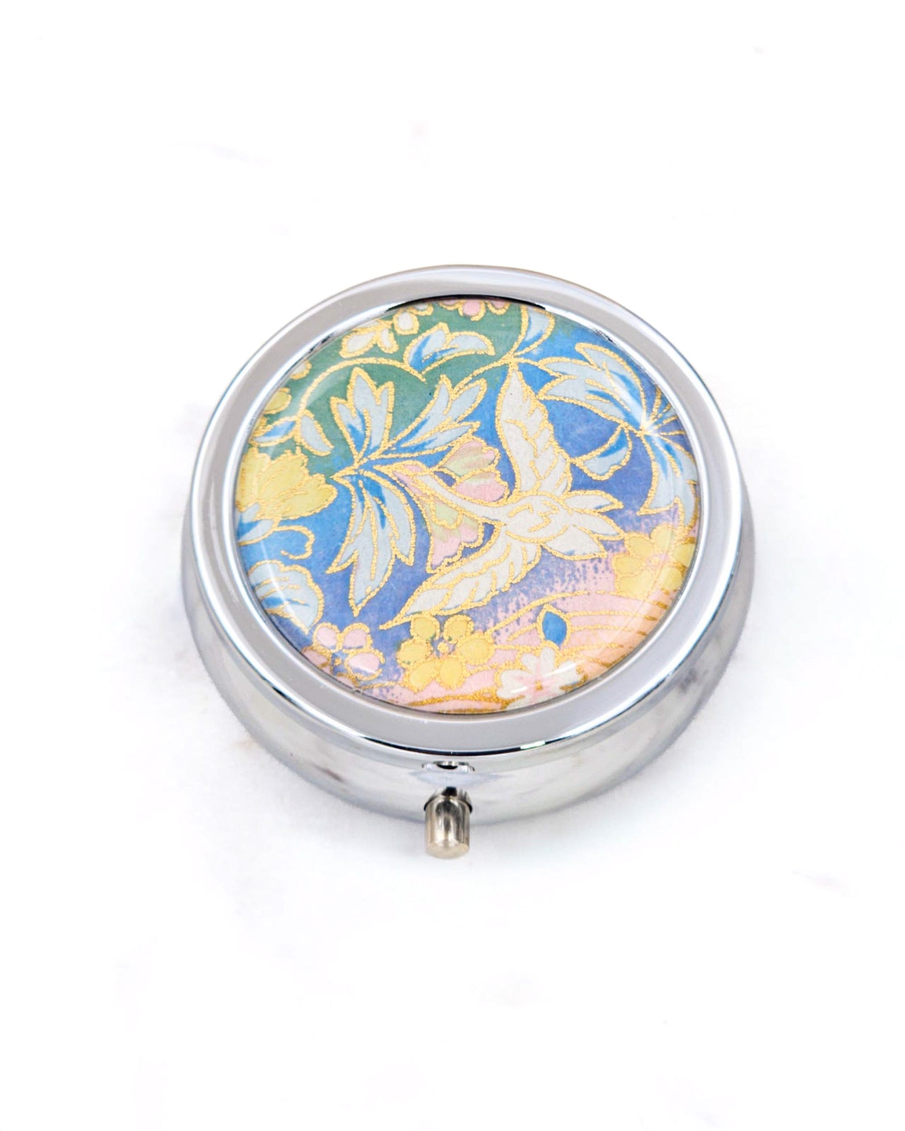 Silver metal pill box featuring a white flying crane on a green, blue , and pink pastel background with metallic gold accents.