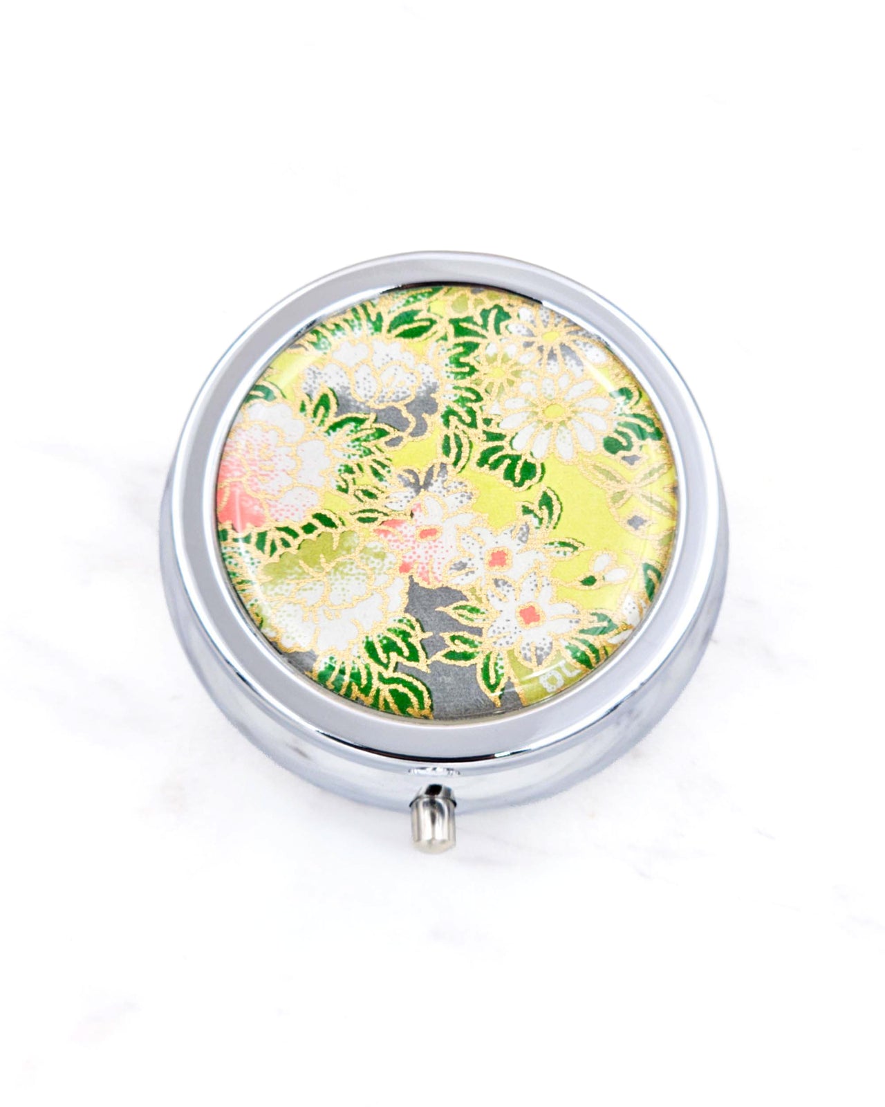 Silver metal pill box featuring assorted white flowers on a green background with metallic gold accents.