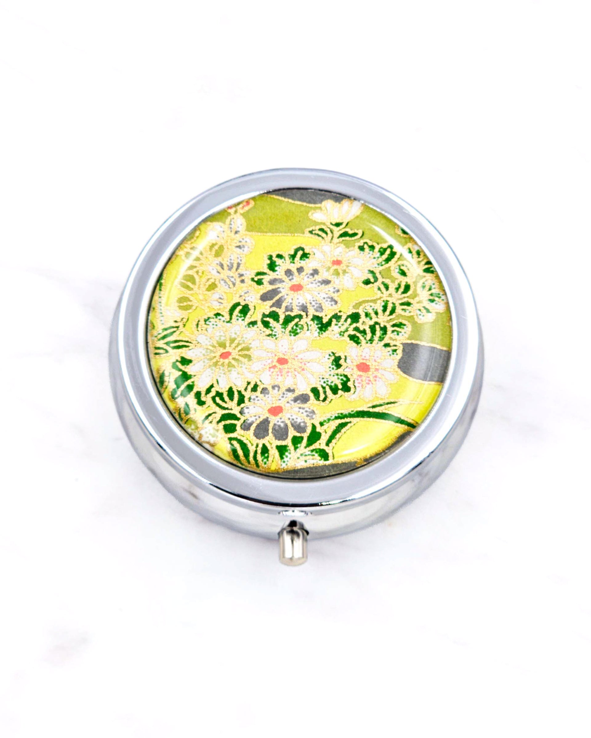 Silver metal pill box featuring white and gray flowers on a green background with metallic gold accents.