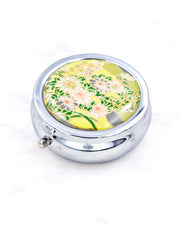 Silver metal pill box featuring white and gray flowers on a green background with metallic gold accents.