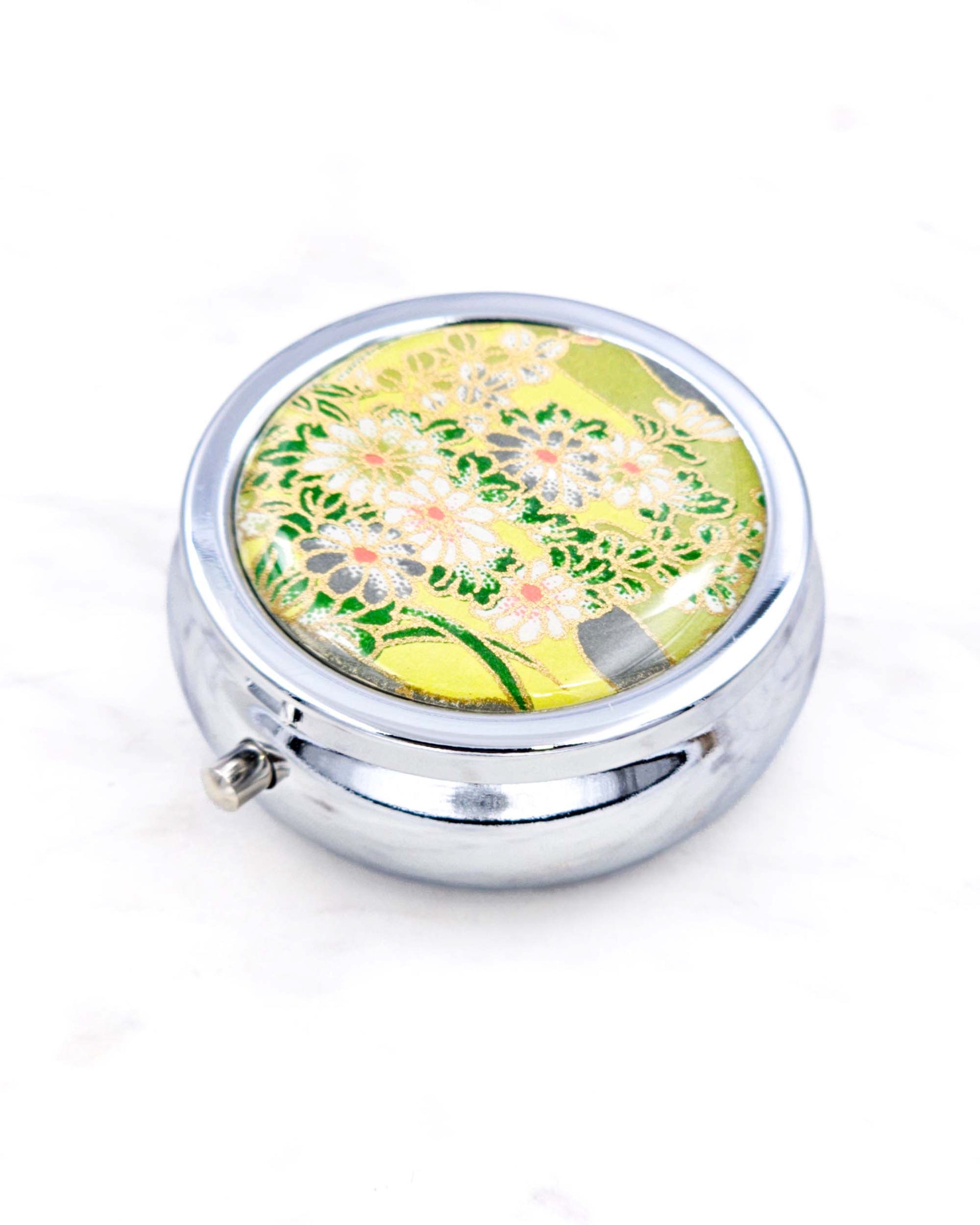 Silver metal pill box featuring white and gray flowers on a green background with metallic gold accents.