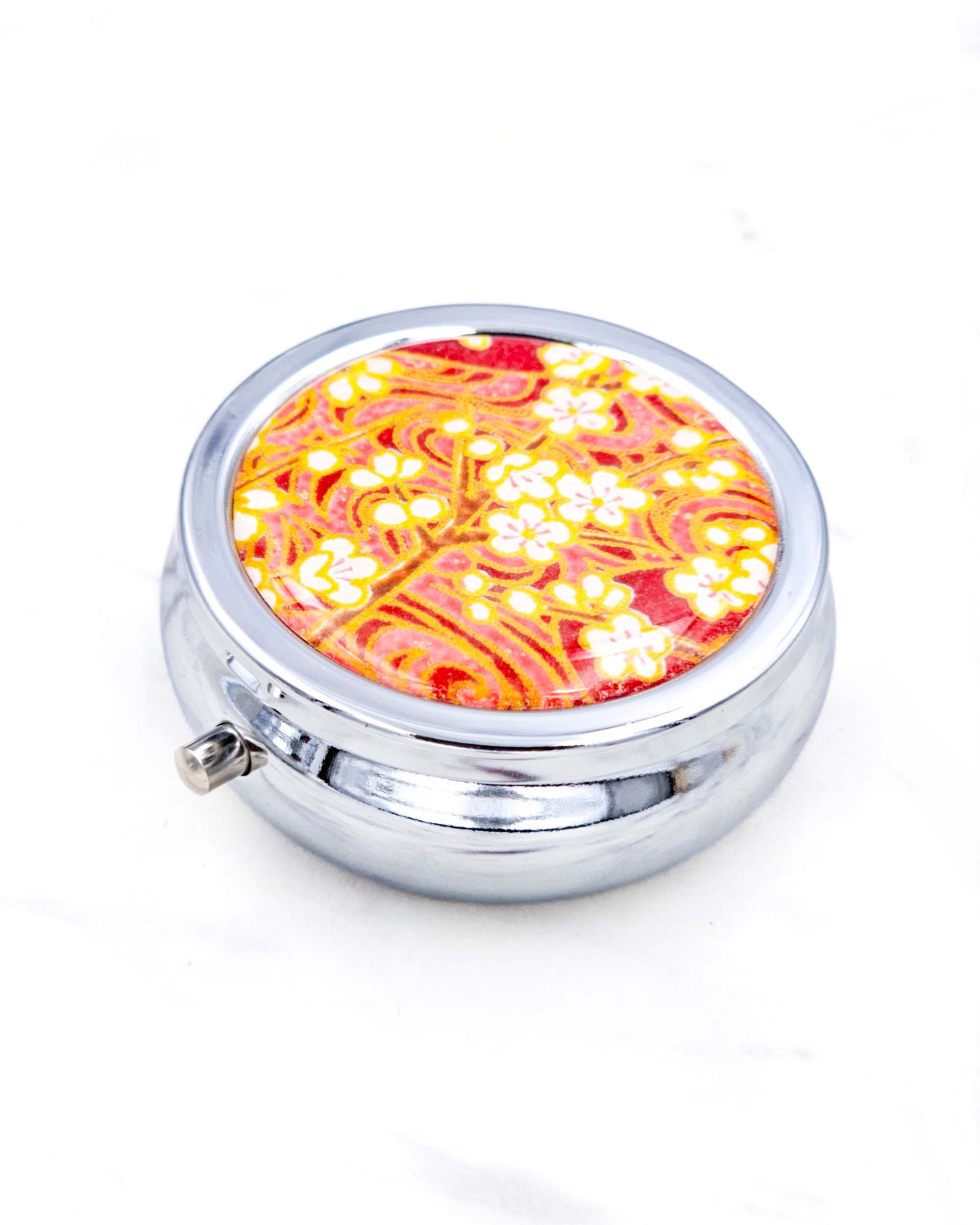 Silver metal pill box featuring white plum blossom branches on a maroon red background with metallic gold accents.