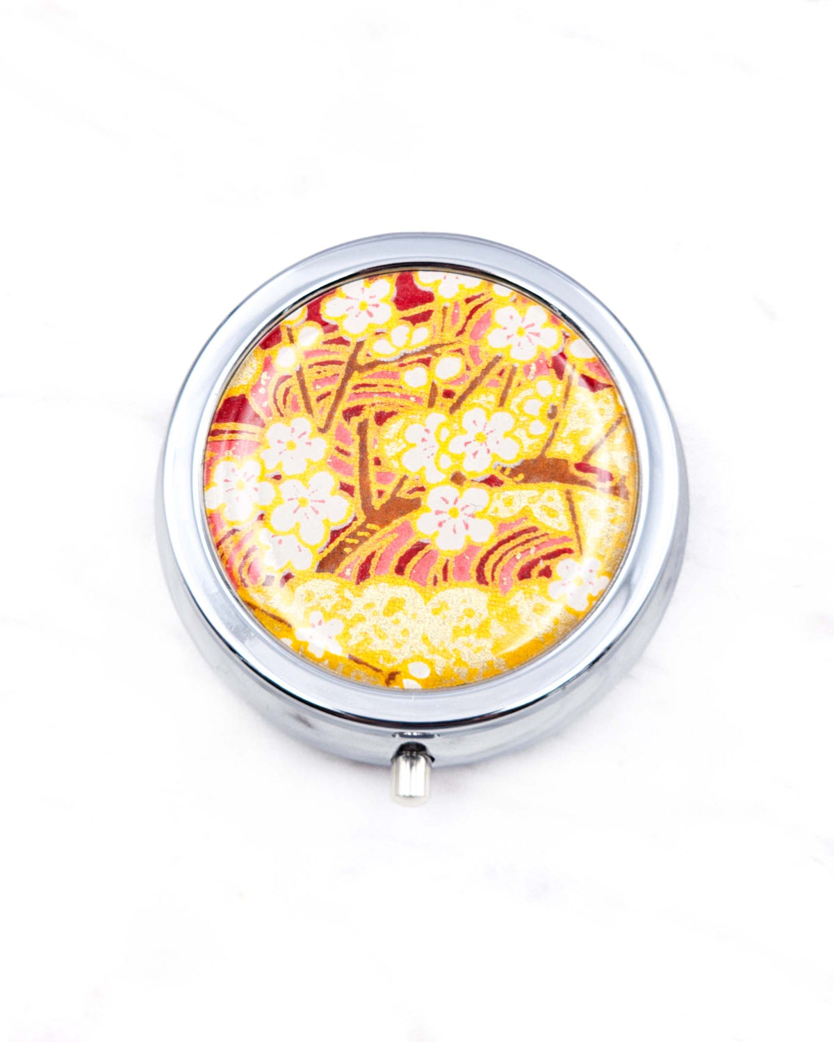 Round silver metal pill box featuring white plum blossom branches on a red and gold background with metallic gold details.