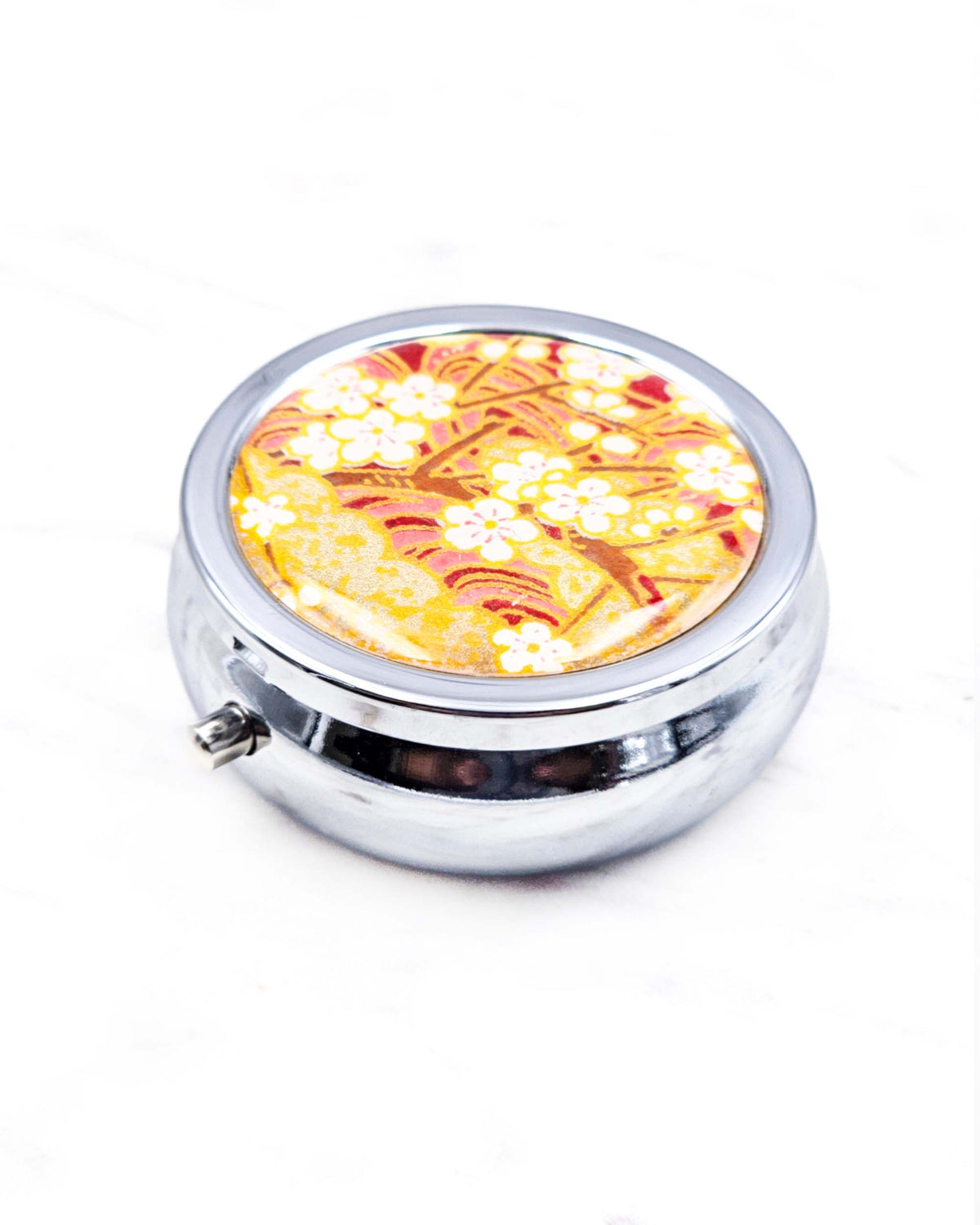 Round silver metal pill box featuring white plum blossom branches on a red and gold background with metallic gold details.