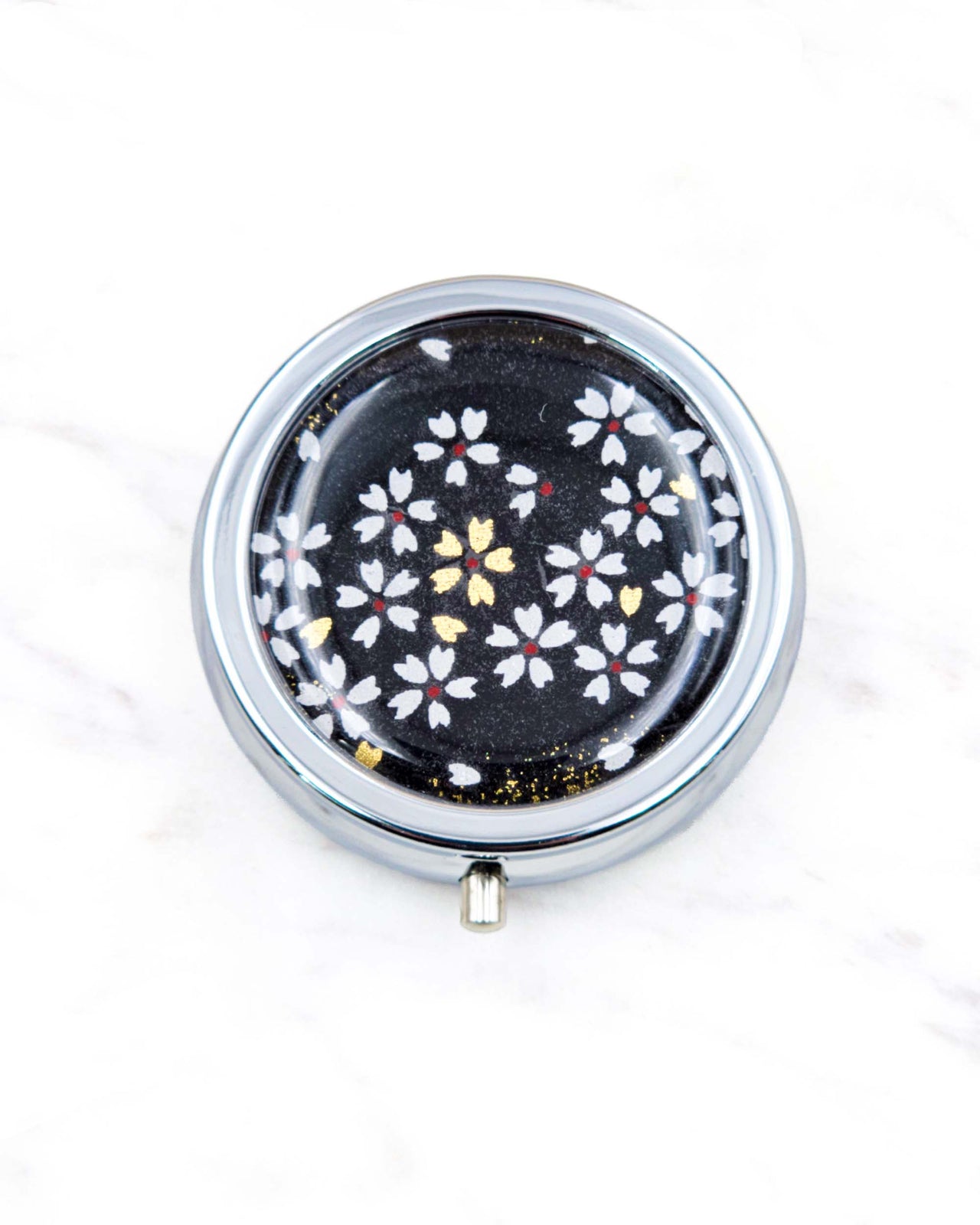 Round silver metal pill box featuring small white and gold cherry blossoms on a black background.