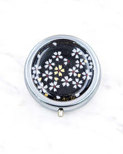 Round silver metal pill box featuring small white and gold cherry blossoms on a black background.