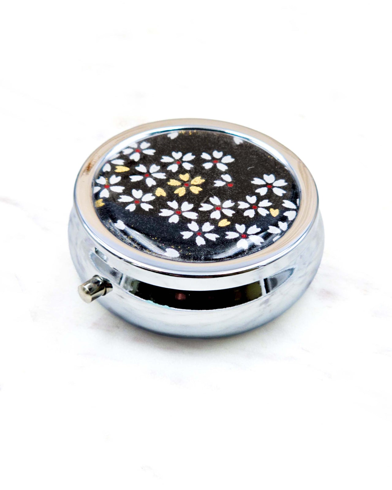 Round silver metal pill box featuring small white and gold cherry blossoms on a black background.