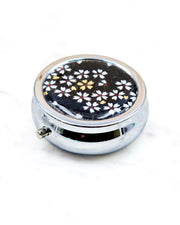 Round silver metal pill box featuring small white and gold cherry blossoms on a black background.