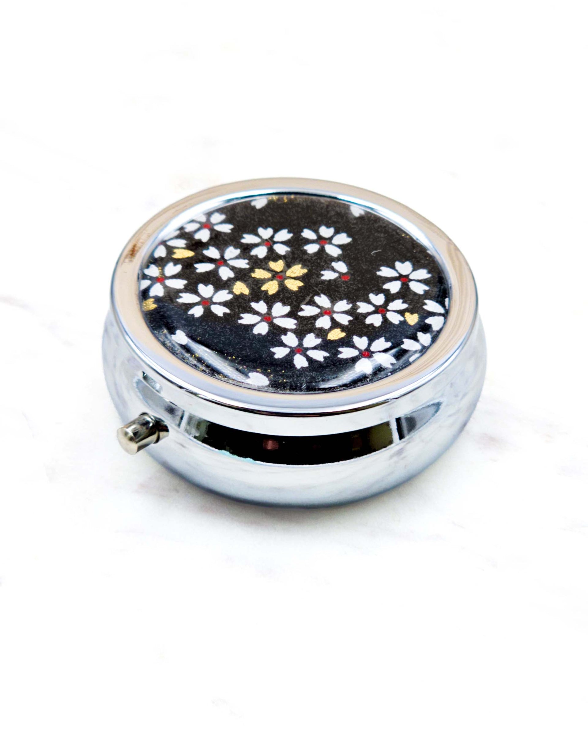 Round silver metal pill box featuring small white and gold cherry blossoms on a black background.