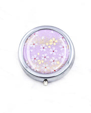 Round silver metal pill box featuring small white and gold cherry blossoms on a purple background with metallic gold details.